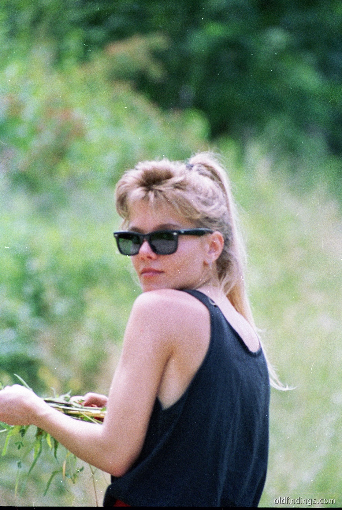 Vintage portrait of a woman in a sleeveless black dress, holding wildflowers in a grassy field. Her retro sunglasses and short, voluminous hairstyle suggest a 1960s–1970s aesthetic. Soft focus and natural lighting enhance nostalgic charm.