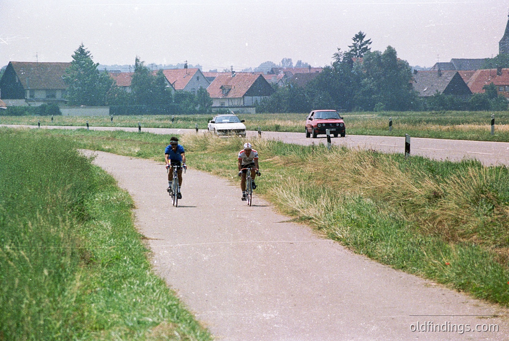 Three cyclists ride along a rural gravel path beside a road with vintage cars (, , ). Traditional Dutch farmhouses with red-tiled roofs line the horizon. Overcast sky and lush greenery suggest a Dutch countryside setting (, , ).