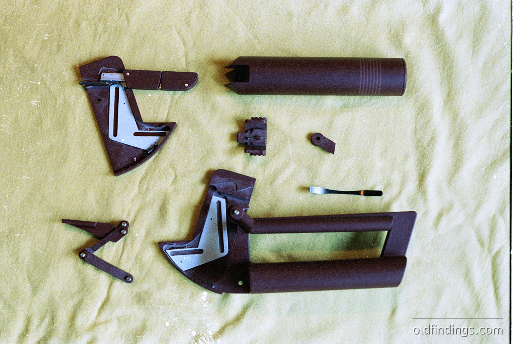 Disassembled vintage metal folding tool set on beige fabric, featuring a cylindrical handle, hinged metal components, and a small brush. Likely a 20th-century utility tool, possibly for mechanical or household use.