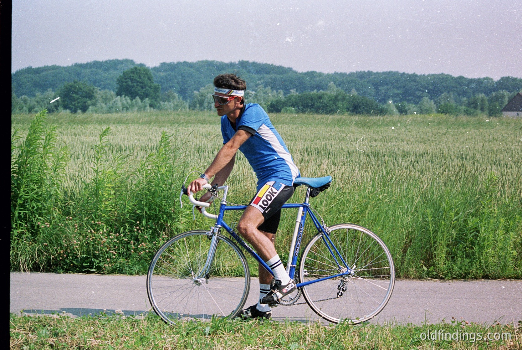 Cyclist in vintage road jersey and helmet rides a blue road bike on a rural road, surrounded by lush green fields and forested hills. The scene evokes 1980s–1990s cycling culture.