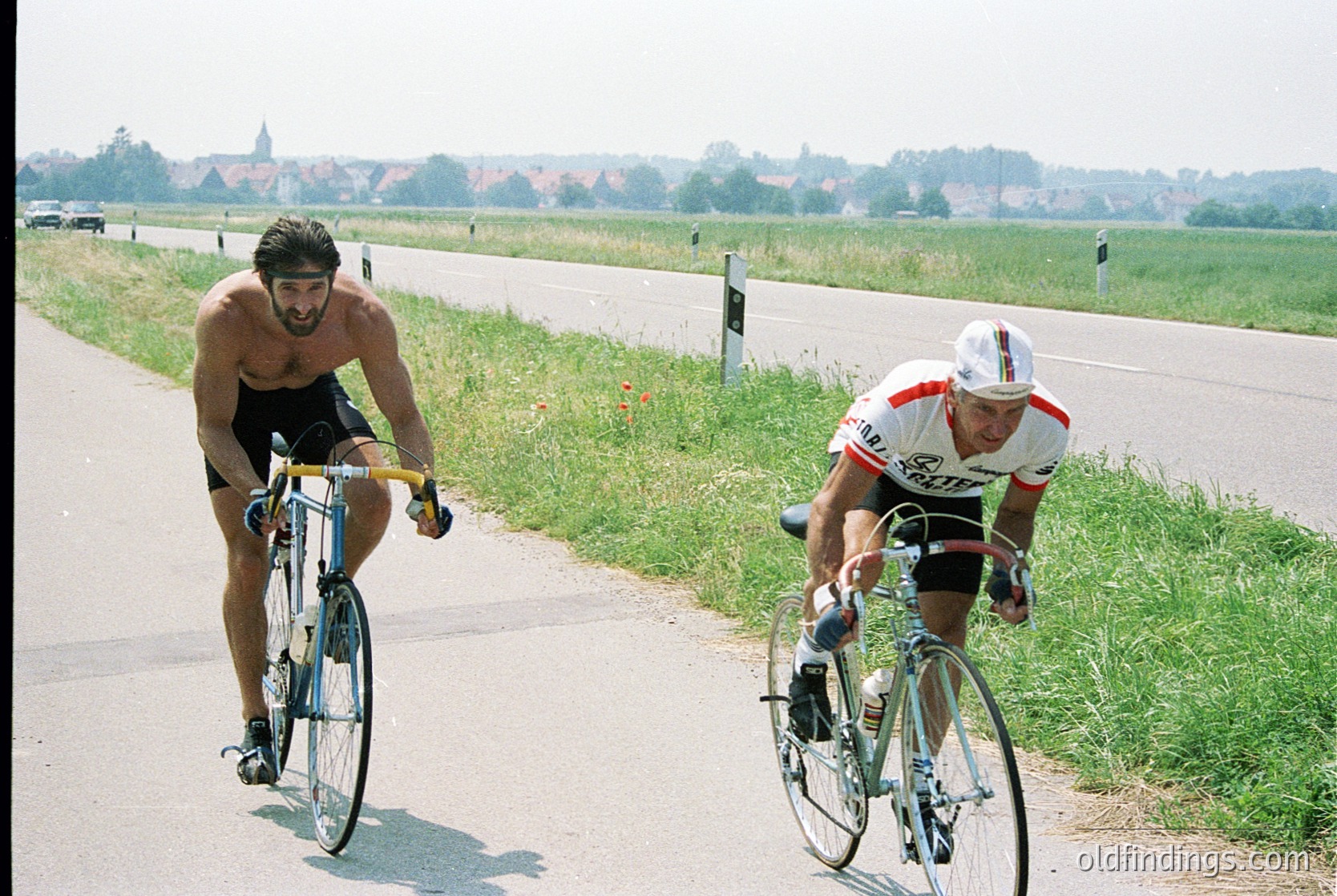 Two cyclists race on a rural road, one shirtless in black shorts, the other in a white/red jersey. Classic road bike frames and drop handlebars suggest 1970s–1980s cycling culture. Open countryside with distant buildings and greenery hints at European countryside. Ideal for sports history, vintage cycling, or retro travel inspiration.