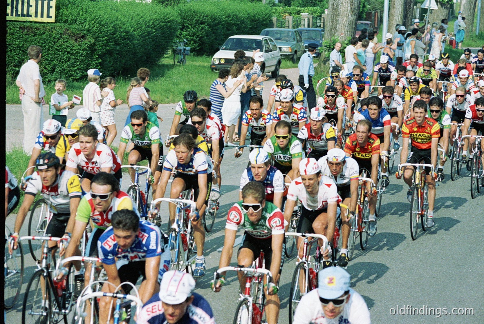Packed cycling race with riders in vintage jerseys and helmets, likely 1970s–1980s. Spectators line the roadside, some in period attire. Dense group dynamics and road racing gear visible.
