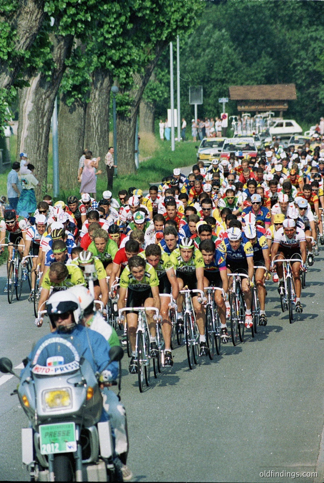 A dense pack of cyclists in vintage racing gear, led by a police escort on motorcycles, races through a tree-lined road. Spectators line the sides, capturing the event. Likely a 1970s–1980s European cycling race ( ).