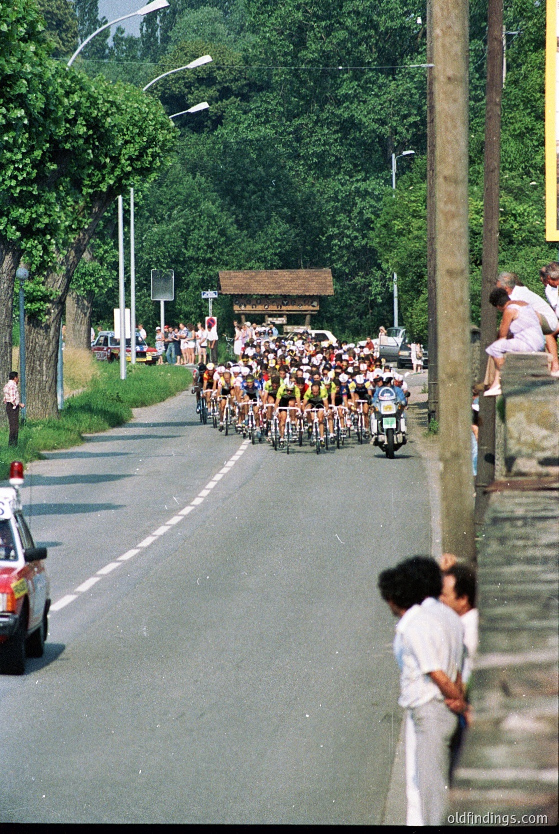 Crowded roadside cycling event with participants on mopeds and bicycles, lined by spectators on both sides. Green trees and a small wooden structure frame the scene, suggesting a rural or suburban setting. Likely a 1970s–1980s European event based on attire and vehicle styles.