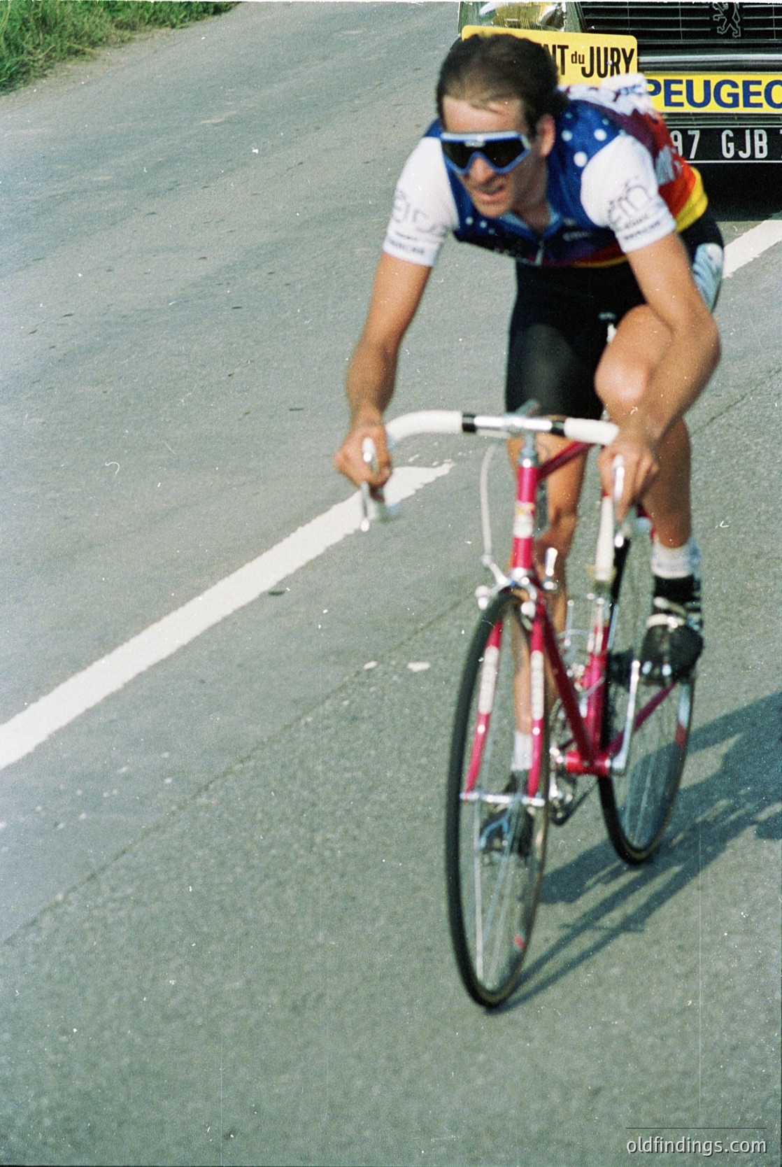 Cyclist in a 1980s road race jersey (Peugeot team) leaning forward on a vintage road bike, gripping handlebars during a sprint. Road markings and sponsor banners (, , , , )