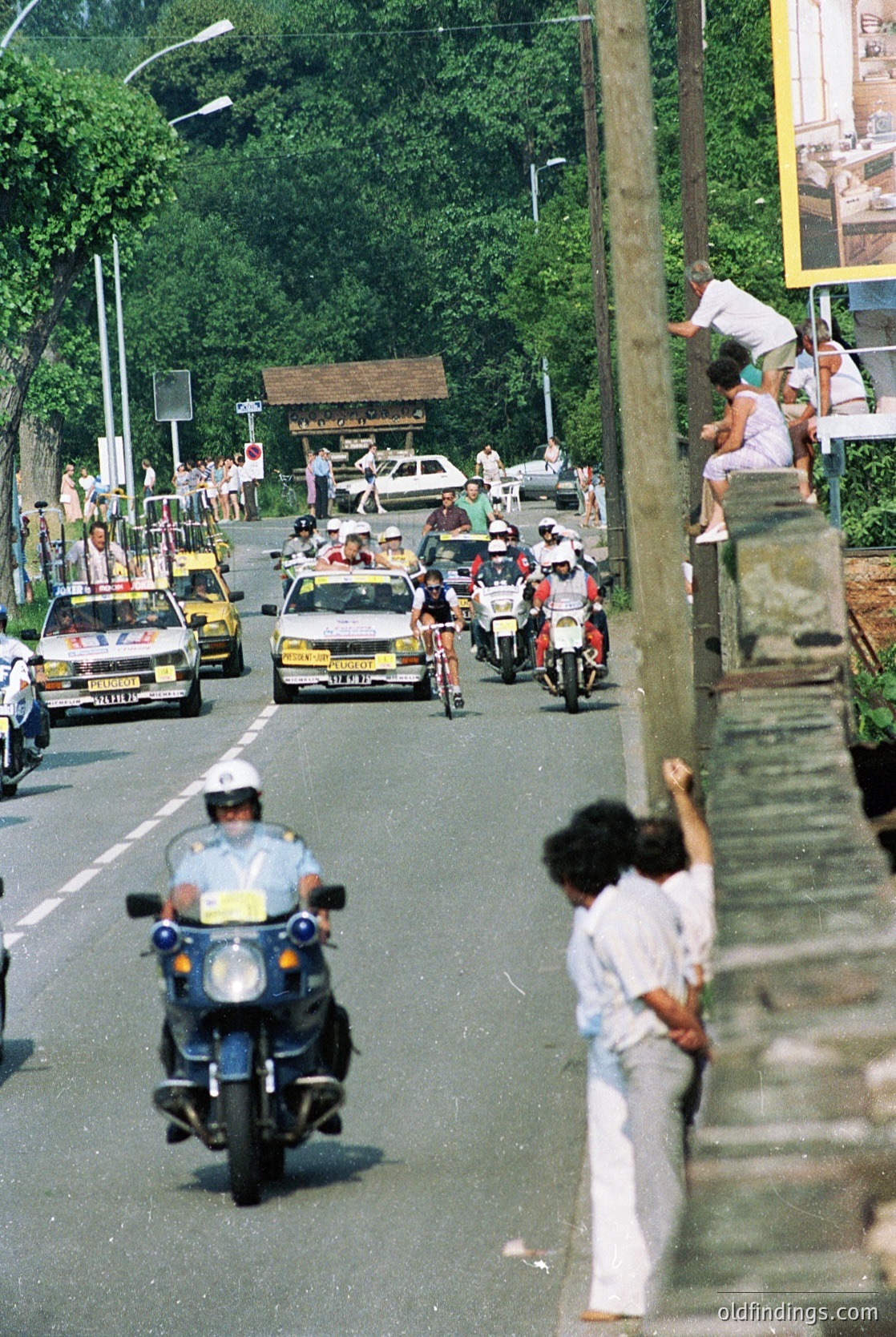 Vintage street scene with motorcade: police motorcycles, vintage cars, and spectators lining a tree-lined road. Mid-20th century urban setting, likely Eastern Europe.