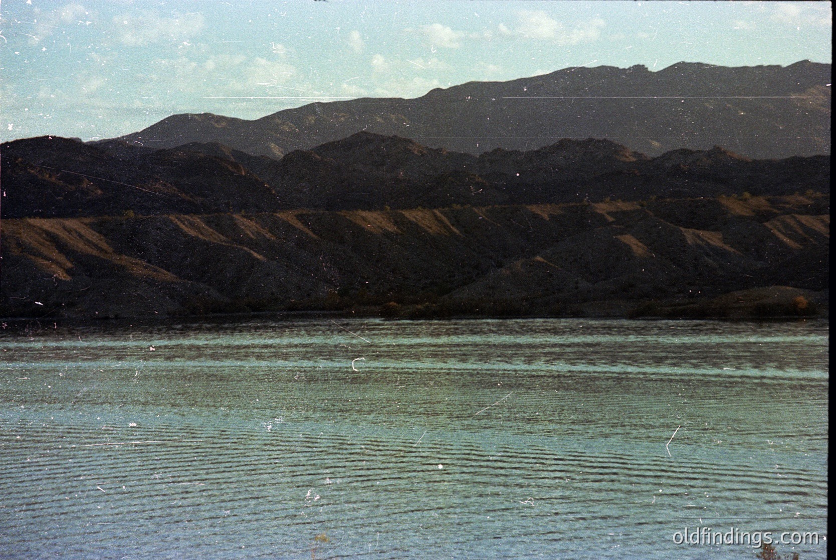 Vintage landscape shot of a serene lake bordered by rugged, forested mountains under a clear sky. The water’s surface reflects muted light, suggesting early/late daylight. Likely captured on film with slight grain, evoking mid-20th century travel or nature photography.