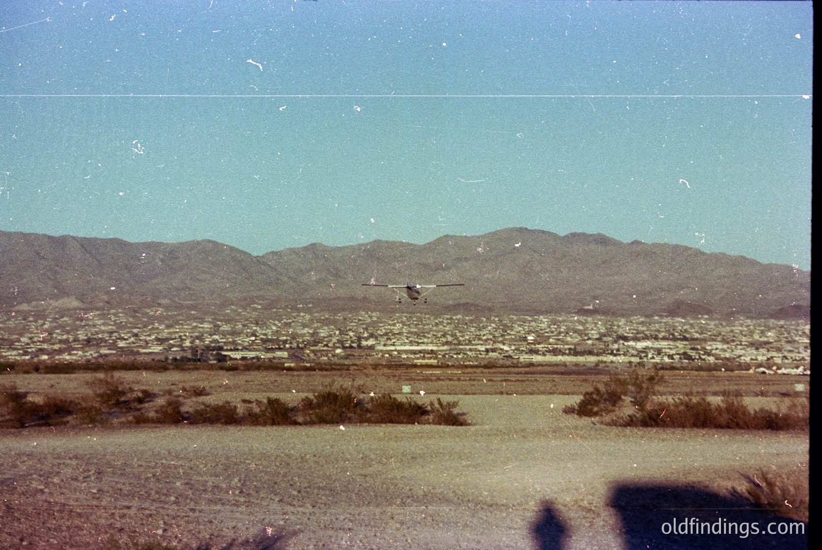 Vintage aerial shot of arid desert terrain with sparse vegetation, likely taken from a small aircraft. Distant mountains frame the horizon, with a single propeller plane in flight. Dusty, low-lying landscape suggests a remote or rural area, possibly the southwestern U.S. or Mexico. *(Note: The image’s grain and lighting suggest a mid-20th-century timeframe, but exact location remains speculative.)*