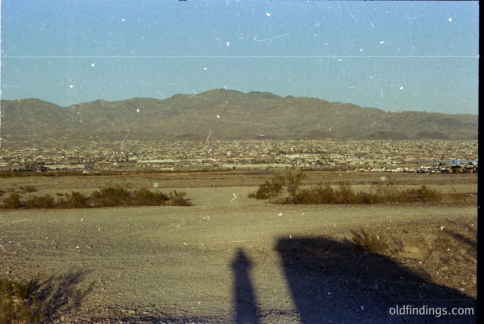 Vintage aerial view of arid landscape with sparse vegetation and distant mountain range. Urban sprawl visible below, suggesting mid-20th century development. Film grain and fading indicate archival quality.