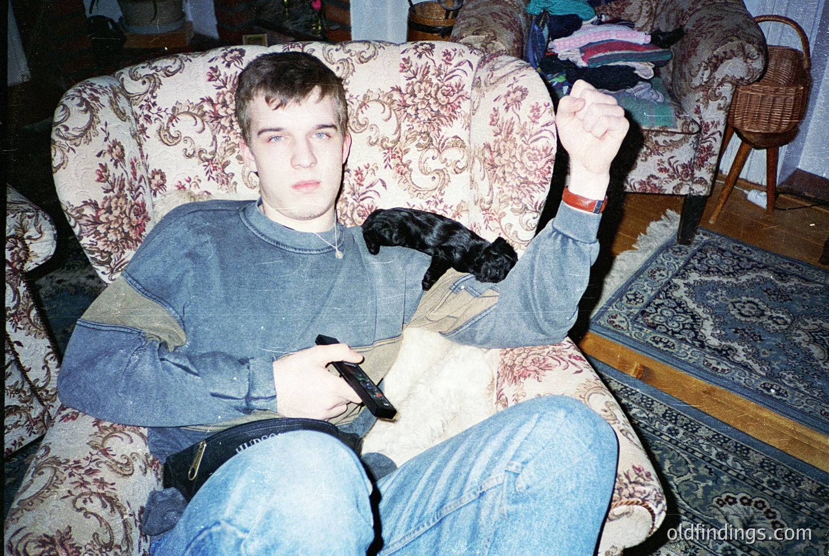 Vintage indoor portrait of a young man playing handheld gaming device (Game Boy) on floral-patterned armchair, mid-1990s. Dog rests on his lap; casual attire includes oversized sweater and jeans. Warm, lived-in interior with wooden floor and woven basket. Evokes nostalgic 90s gaming culture.