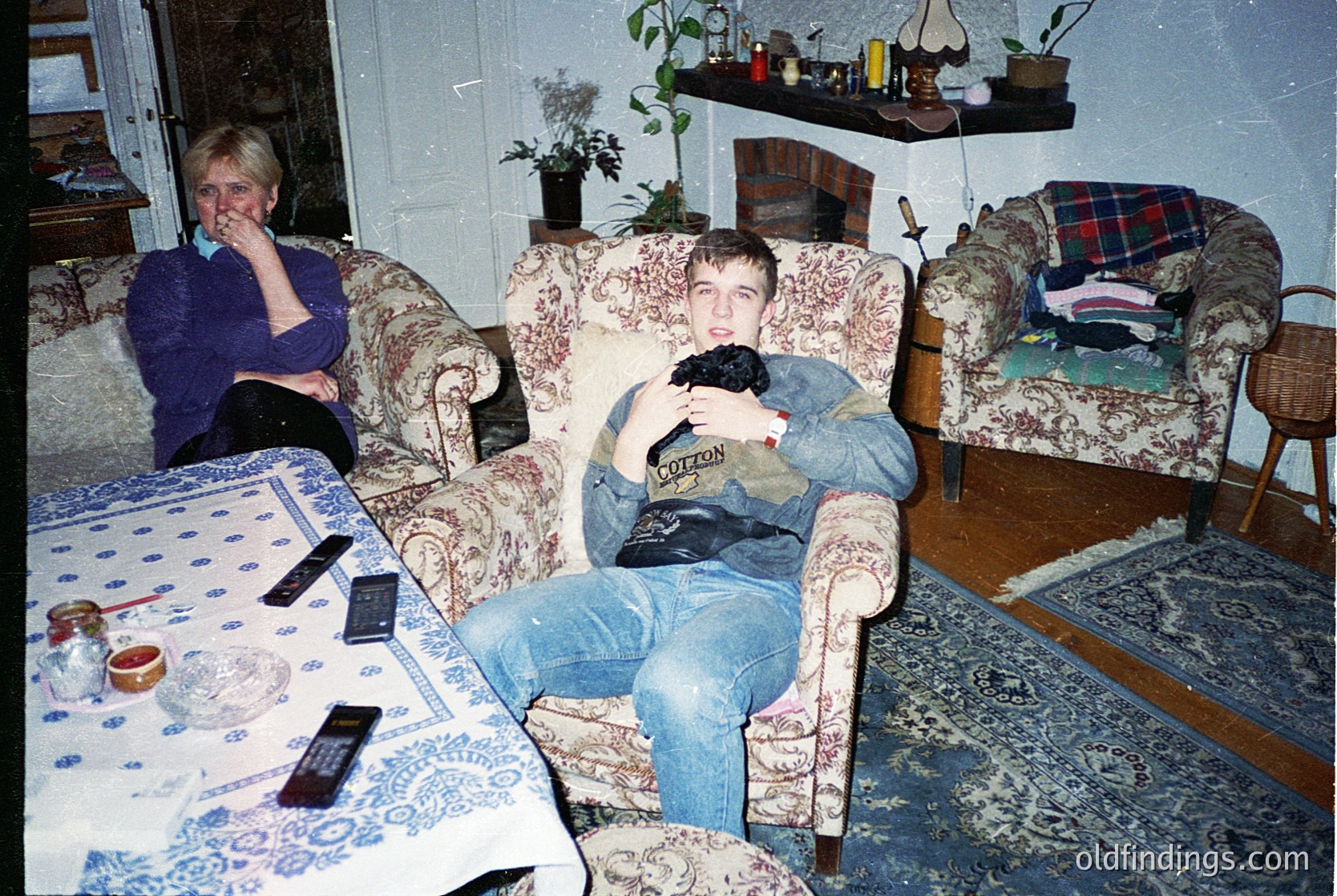 Vintage living room scene with two individuals in mid-1990s/early 2000s décor. Woman in blue sweater rests on floral-patterned armchair beside a table with blue-and-white patterned tablecloth. Man in denim and hoodie poses with a toy gun on a matching floral sofa. Decor includes a fireplace shelf with plants, a checkered blanket, and a wooden rocking chair.
