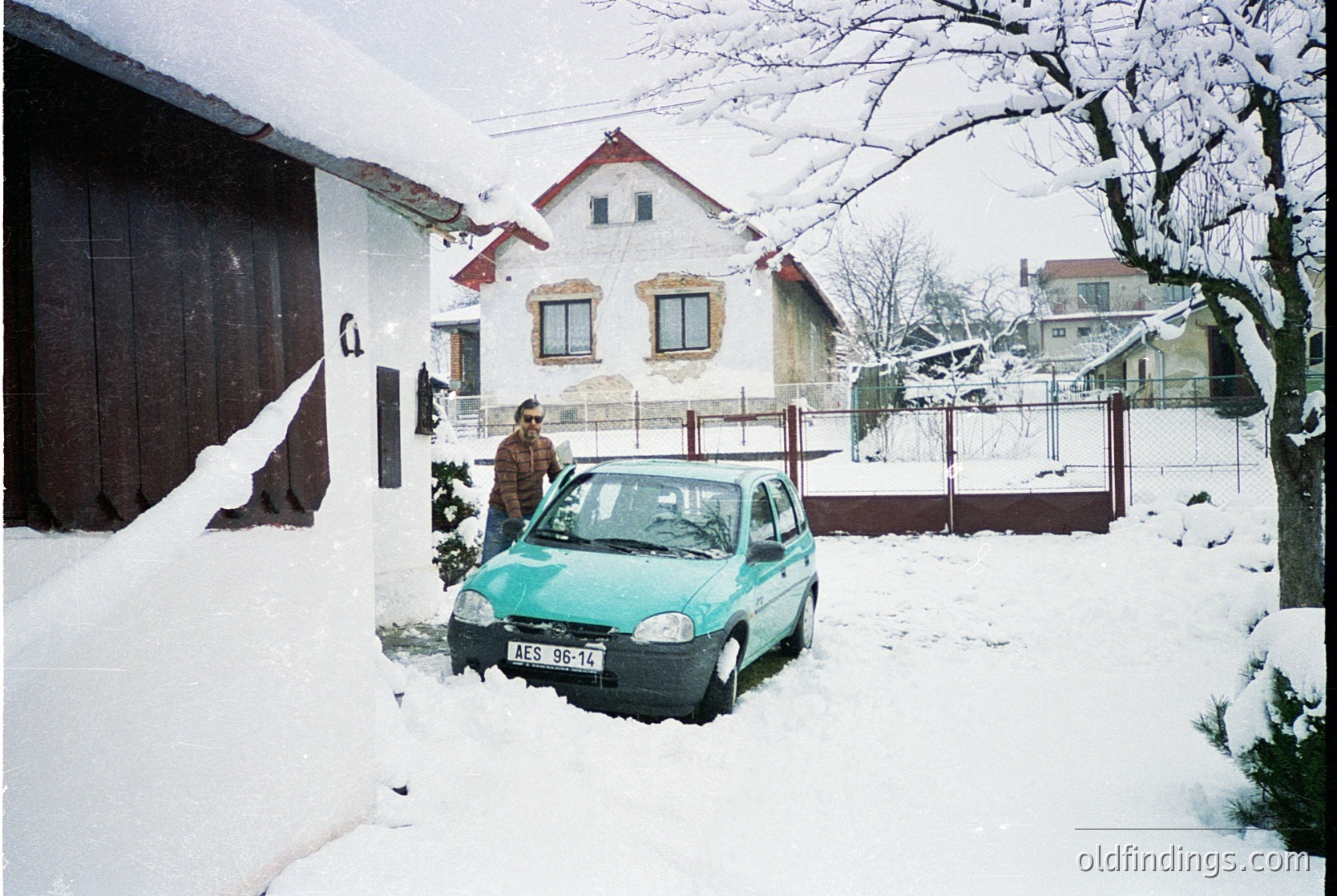 Snow-covered residential street with 1990s-era compact car (turquoise, likely Škoda) parked beside a light-colored house. Man in winter coat stands near the vehicle, while a barn-style garage dominates the left. Snow blankets rooftops, fences, and ground, indicating heavy winter conditions. Urban/suburban setting, likely Eastern Europe.