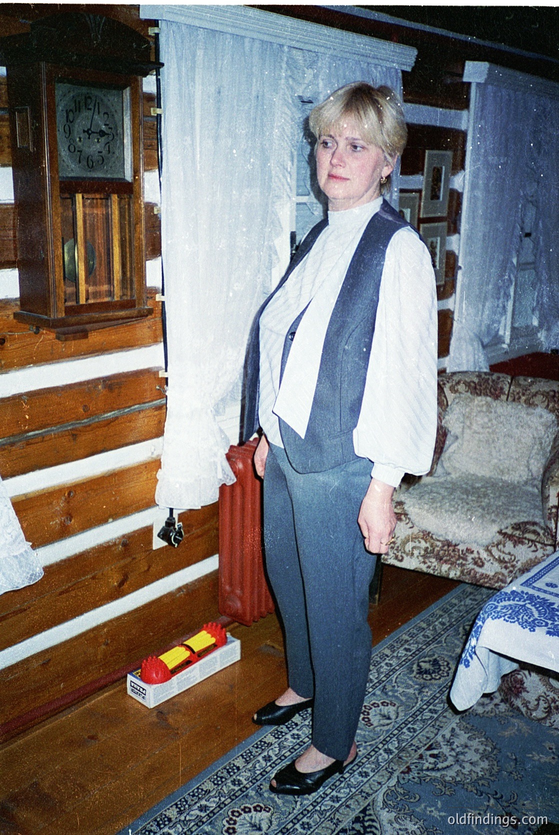 Vintage interior featuring a woman in a 1970s-style blazer and high-waisted trousers, posing beside a wooden-framed clock and patterned rug. Rustic wooden beams and a floral-patterned sofa add warmth. Red suitcase suggests travel or relocation.