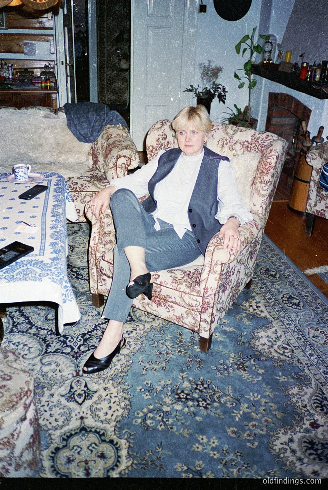 Vintage interior portrait of a woman in a relaxed pose on a floral-patterned armchair, wearing a blazer over a white top, high-waisted jeans, and black tights with loafers. Surroundings include a patterned rug, a table with a blue-and-white tablecloth, and a shelf with decorative items. Likely 1970s–1980s Western home decor.