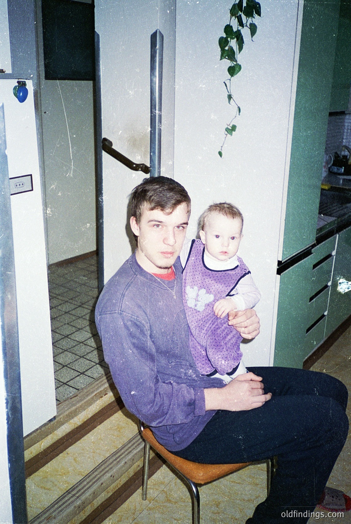 A man in a purple sweater holds a toddler in a matching vest on a wooden chair in a utilitarian indoor setting. White-tiled floor and green-painted walls with exposed wiring suggest a mid-20th-century residential space. [Vintage family portrait in Soviet-era apartment ]