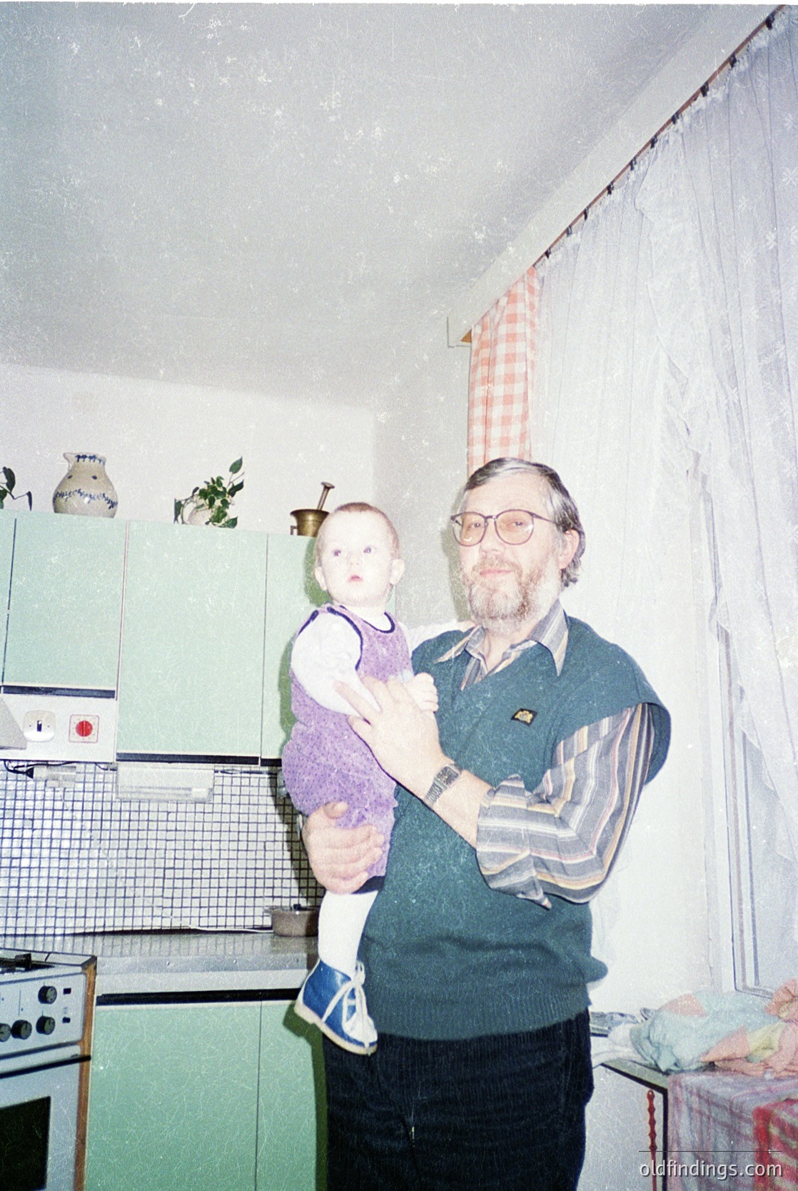Mid-20th century kitchen interior featuring a man in a striped sweater and glasses holding a baby in a purple sweater and blue sandals. Green metal cabinetry and a white stove with a clock atop it. Checkered curtain and plastic shelf decor. Likely Eastern Bloc-era domestic life.