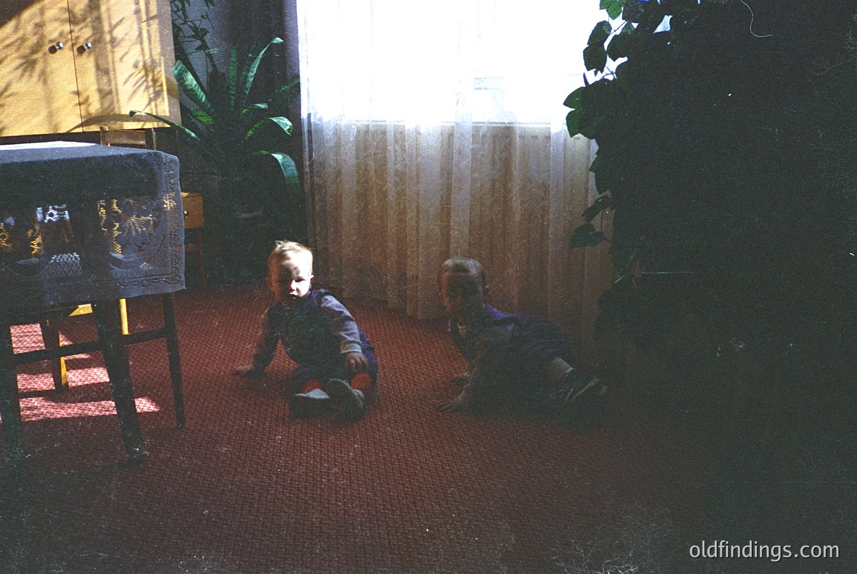 Vintage indoor scene of two young children playing on a red carpeted floor, likely mid-20th century. Child on left sits near a wooden table with a patterned cloth, while the other crawls toward a striped curtain. Warm lighting and greenery add homely warmth.