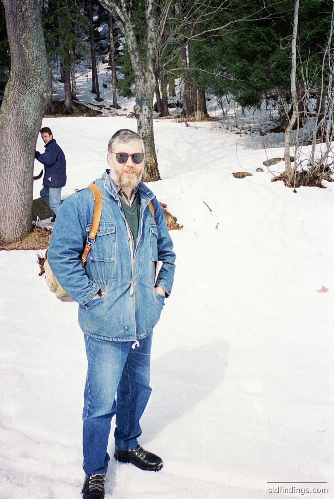 Man in vintage denim jacket and sunglasses poses on snow-covered trail, carrying a crossbody bag. Leather boots and bare patches of ground suggest early spring. Second person in dark jacket and jeans in background.