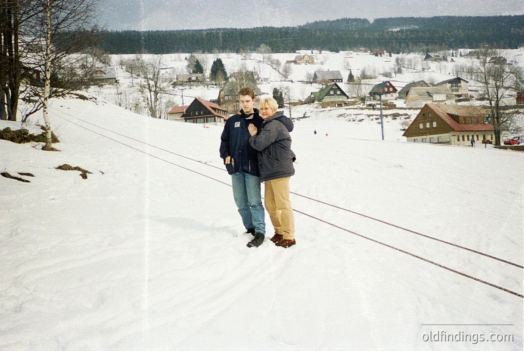 Two individuals embrace on a snow-covered slope beside a railway track, surrounded by a quaint village blanketed in winter. The vintage aesthetic suggests a mid-20th-century European alpine setting.