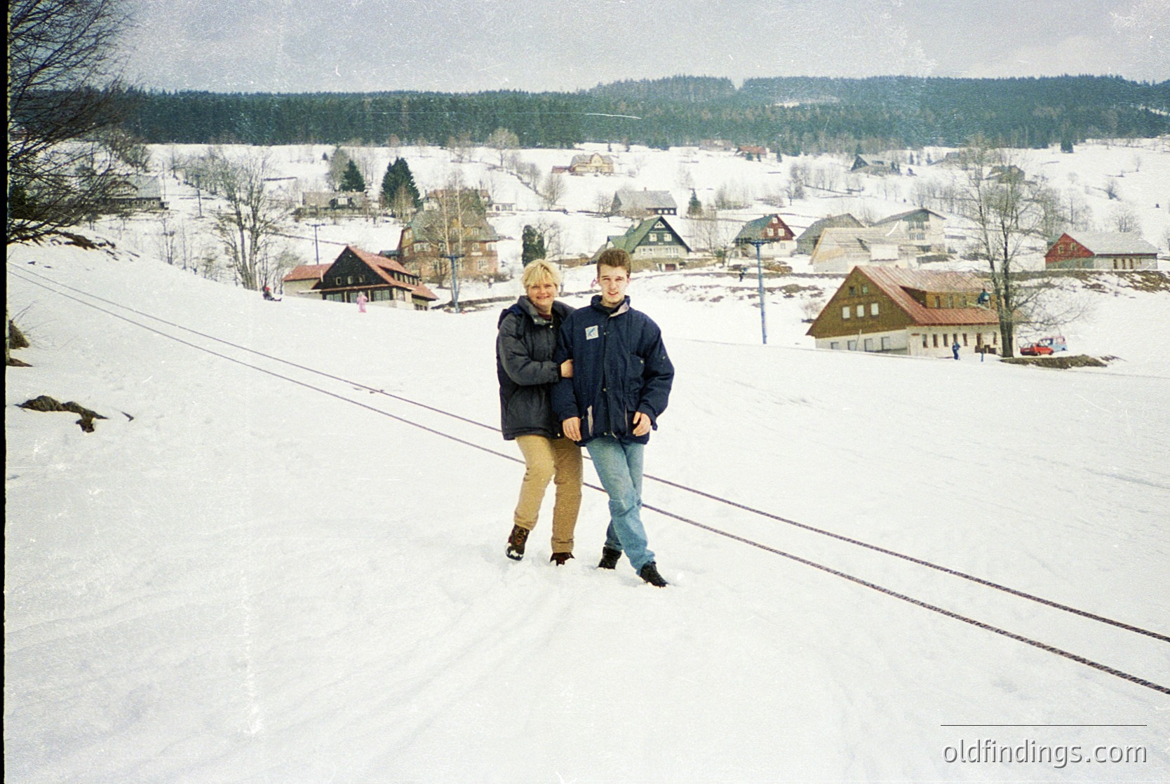Two individuals pose on a snow-covered railway track in a winter village, surrounded by snow-dusted buildings and evergreen forests. The scene suggests a 1980s-1990s European alpine setting, likely for nostalgic or travel inspiration.