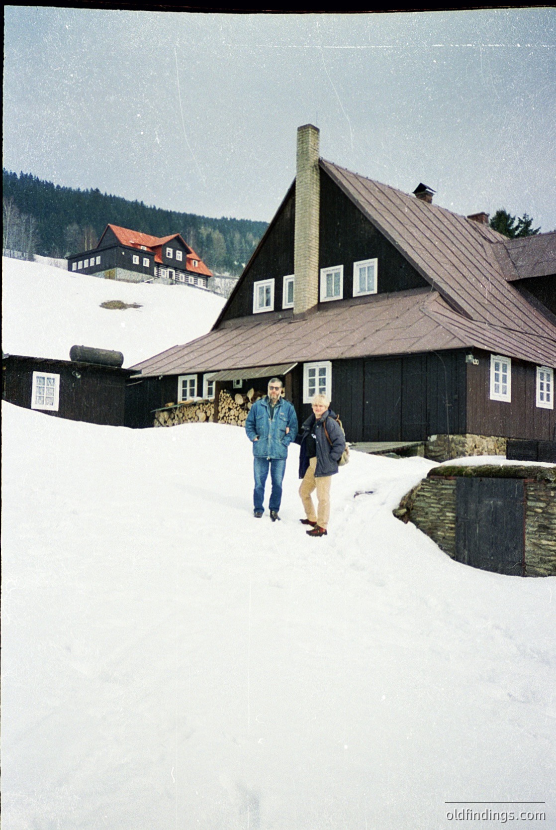 Two men in winter attire pose in front of a dark wooden alpine chalet with a steep gabled roof, surrounded by snow-covered terrain. Stacked firewood and a chimney indicate a rustic, mountainous setting. Likely 1960s–1980s European Alps.