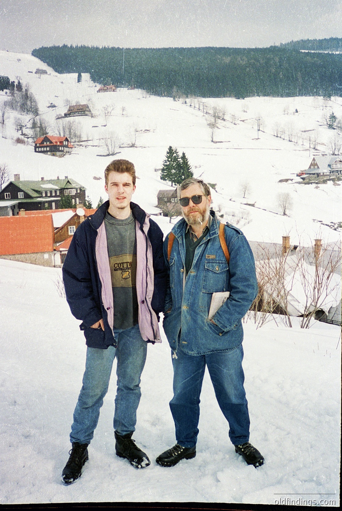 Two men pose in winter attire on a snowy hillside overlooking a quaint village. The younger wears a purple scarf, dark jacket, and jeans; the older sports sunglasses, a denim jacket, and a backpack. Snow-covered rooftops and evergreen trees frame the scene, suggesting a European alpine village. Likely late 20th century (1970s–1990s).