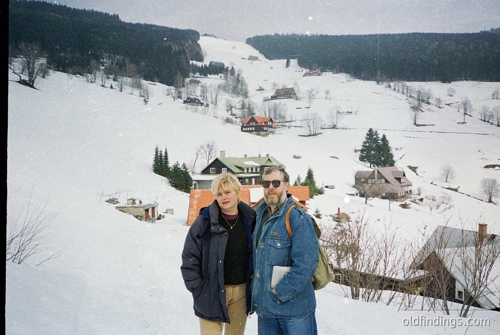 Two individuals pose on a snow-covered slope in a winter village, surrounded by wooden chalets with red roofs. The man wears a denim jacket, sunglasses, and a backpack; the woman wears a dark coat and beige pants. Snow-capped hills and evergreen trees frame the scene, suggesting a European alpine setting. Likely late 20th century (1970s–1990s).