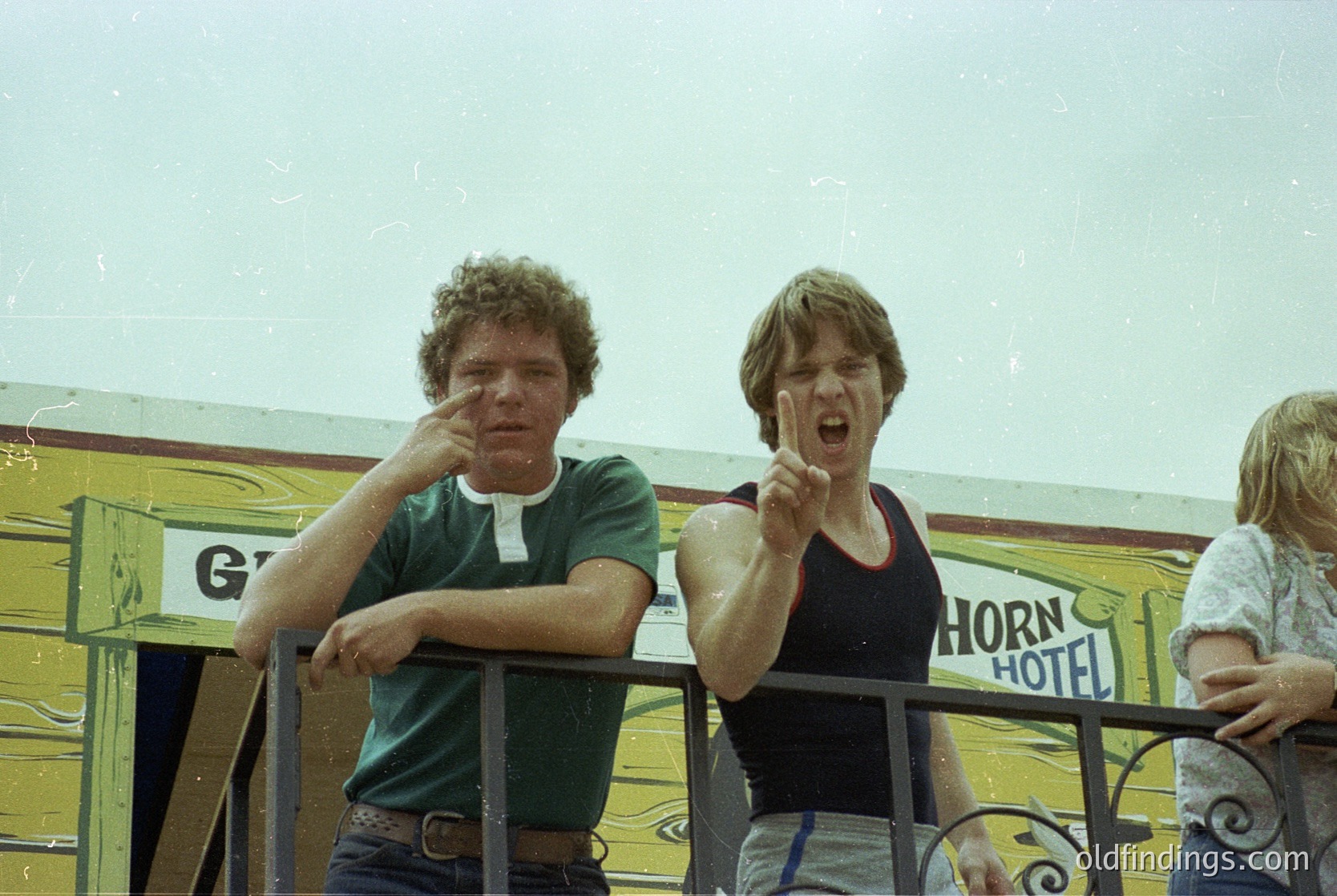 Vintage photo of two men posing on a vintage "Horn Hotel" bus, likely 1970s. One wears a green shirt, the other a black tank top. Bus features retro signage and ornate railings. Candid, expressive gestures capture youthful energy.