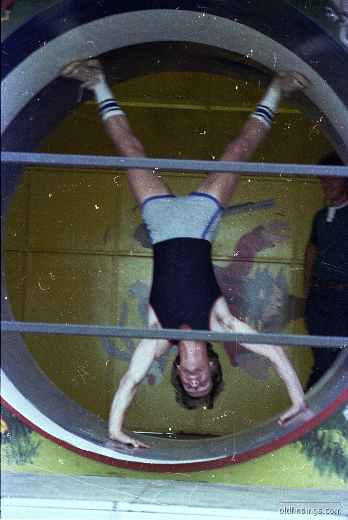 Upside-down gymnastics routine on a horizontal bar, likely in a vintage gymnasium. The athlete wears a striped sleeveless top and black shorts, gripping the bar with both hands. Reflective surface behind suggests polished concrete or tiled floor.