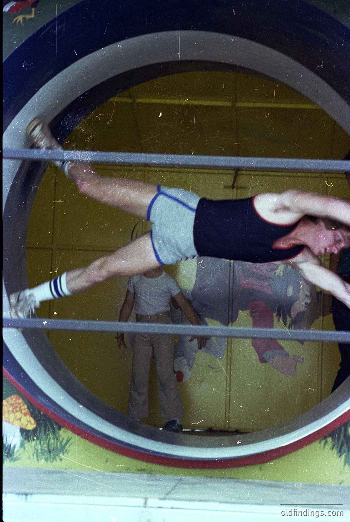 Vintage gymnastic scene through a circular window frame, showing a male athlete mid-dynamic move on parallel bars. White shorts with blue stripes and white wristbands visible. Reflective glass distorts background—likely a 1960s–1970s Soviet-era sports hall.