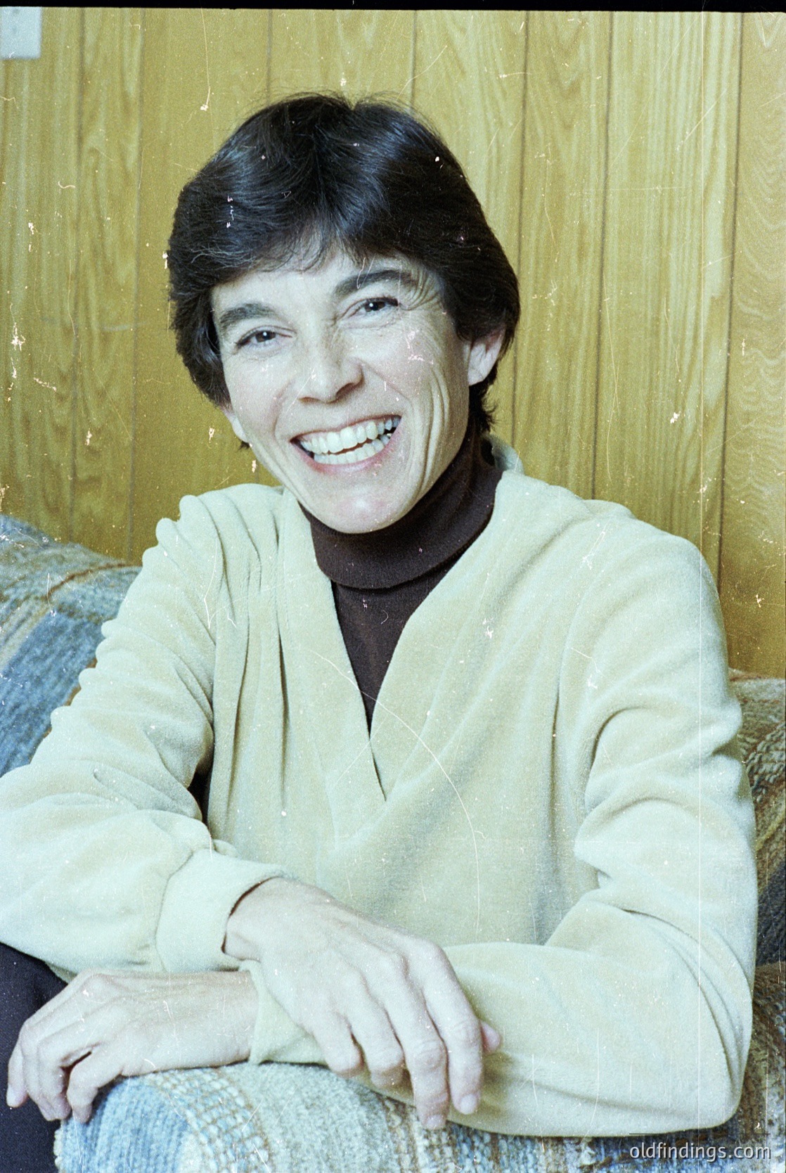 Portrait of a smiling individual in a light-colored, long-sleeve sweater with a V-neck, seated indoors against a wooden-paneled wall. The setting suggests a mid-20th century domestic interior, likely 1960s–1970s. Warm lighting enhances the candid, relaxed expression.