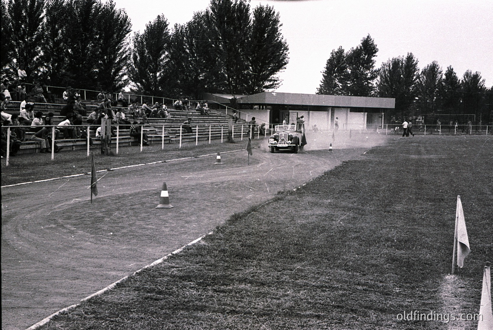 Vintage black-and-white shot of a dirt track racing event, featuring a lone driver in a classic open-wheel car navigating a turn. Spectators line tiered wooden bleachers under shaded trees. Small orange traffic cone and flag mark track boundaries. Mid-century concrete grandstand and simple clubhouse in background.