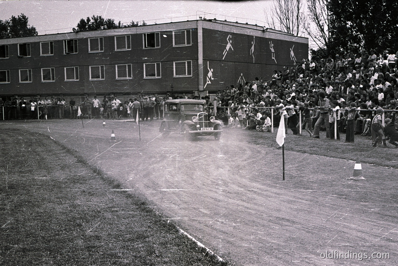 Vintage black-and-white shot of a mid-20th-century auto race on a dirt track. A vintage car speeds past spectators lined behind barriers, with a multi-story building featuring mural-style murals of athletes in the background. Crowds gather along the trackside, suggesting a community event or local competition.