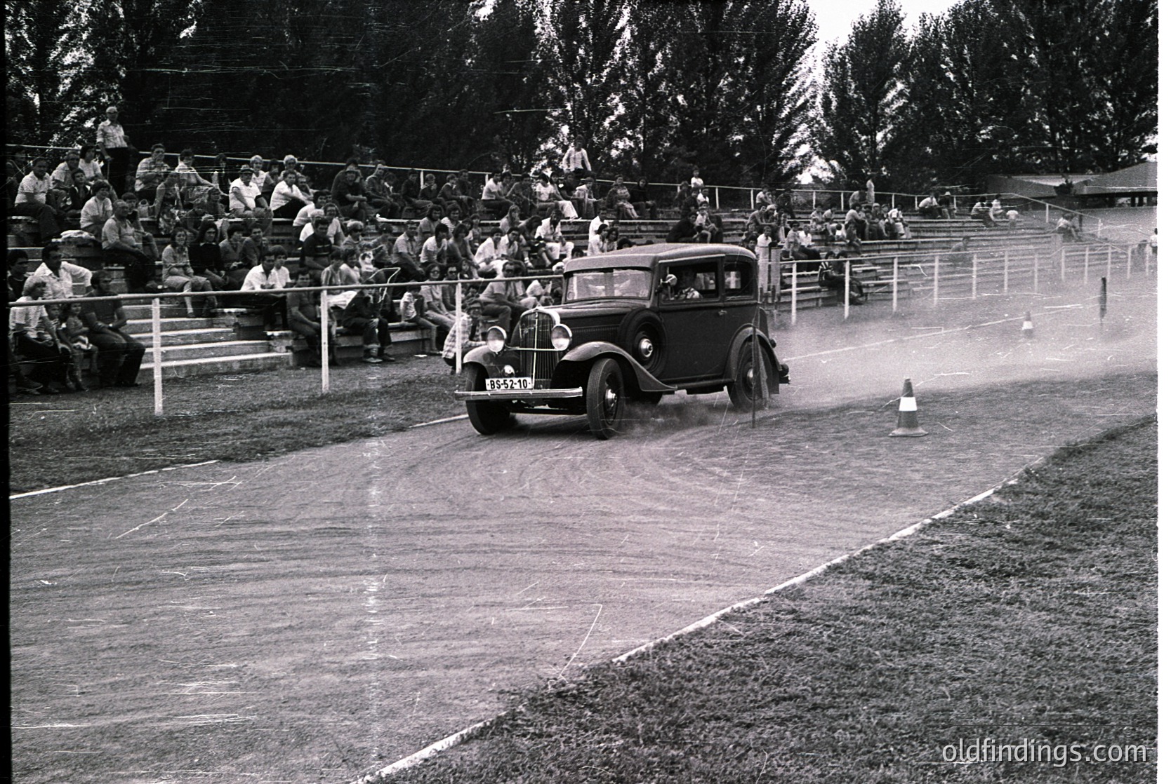 Vintage black-and-white photo of a classic 1930s-era sedan navigating a muddy track during a slalom race. Spectators line wooden bleachers, some holding umbrellas against rain. Wet surface and spray highlight the event’s challenging conditions.