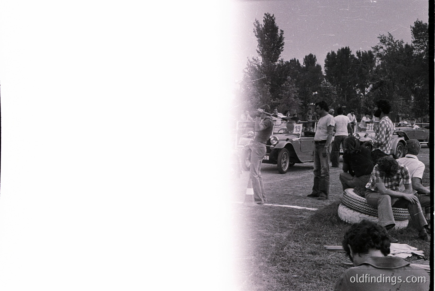 Vintage black-and-white photo of a 1960s outdoor gathering. A vintage car (likely a Volkswagen Beetle) with a white roof and red accents drives past a crowd of casually dressed people, some seated on grass. Trees and a fence frame the scene, suggesting a rural or suburban setting.
