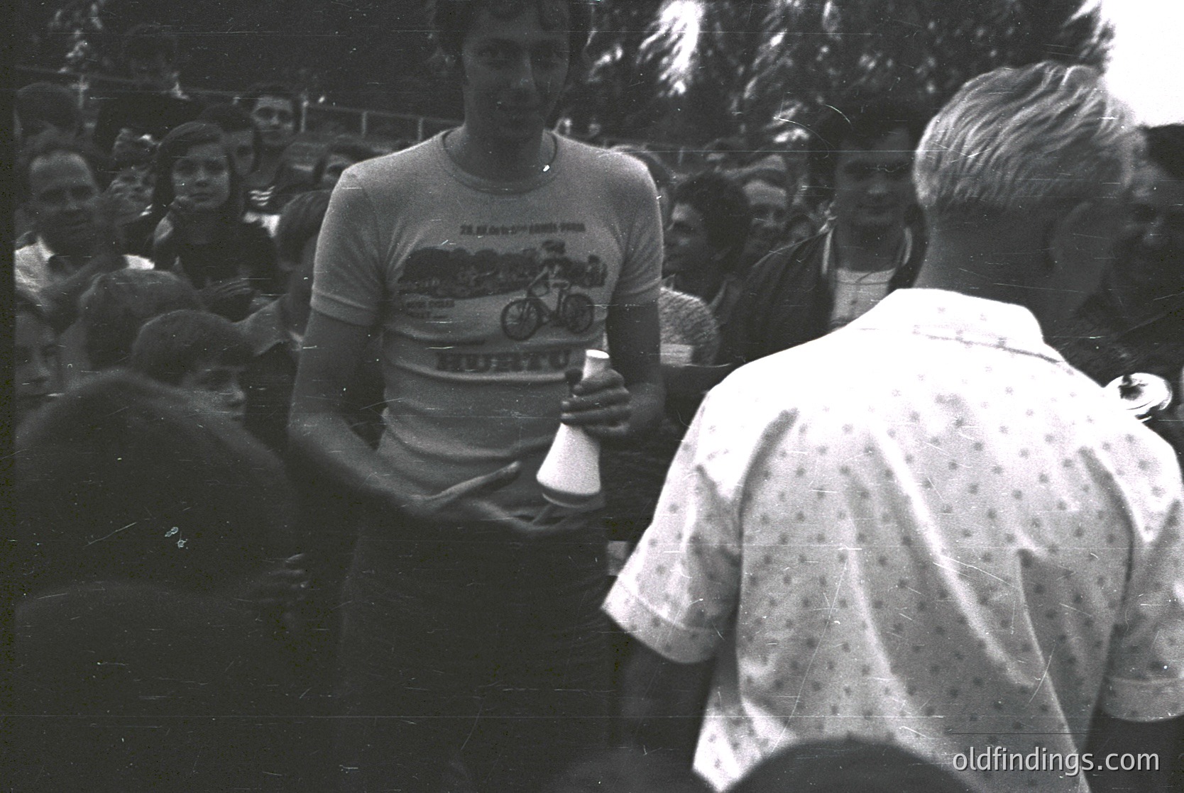 Cyclist in 1970s racing jersey receiving trophy from official at outdoor event. Crowd in background, suggesting post-race celebration or podium moment. Classic retro sportswear and vintage photography style.