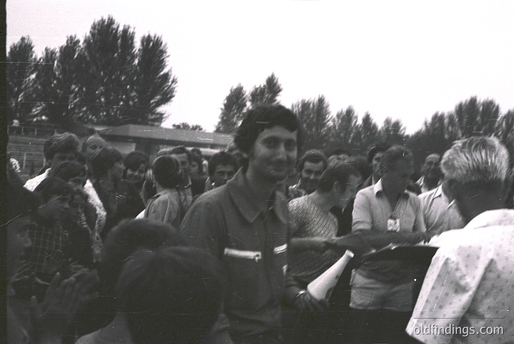 Vintage black-and-white photo of a crowded outdoor gathering, likely a public event or rally. Central figure in a striped shirt shakes hands with a man in a light-colored shirt. Casual attire suggests mid-20th century (1950s–1970s). Wooden fence and trees in background indicate rural or suburban setting. Crowd appears engaged, suggesting political or community significance.