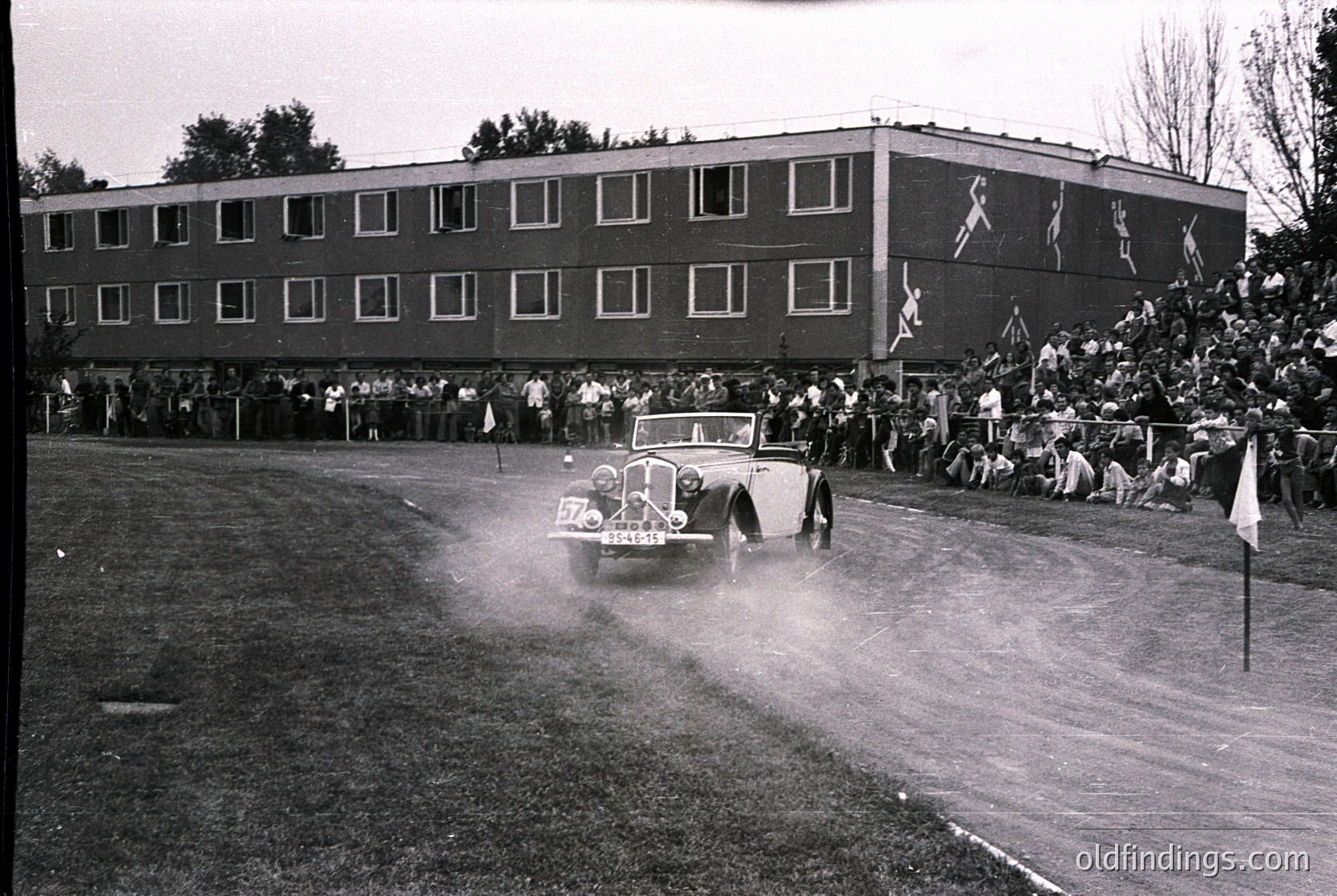 Vintage black-and-white photo of a classic car race on a wet track, 1950s. A vintage convertible (likely a pre-war model) speeds through spray, numbered 570. Spectators line a multi-story brick stadium with mural-style murals on the facade. Crowds behind barricades, some holding flags.