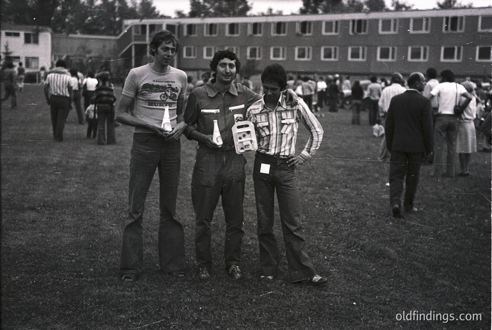 Three men pose outdoors in 1970s-era attire—striped polo shirts, rolled-up sleeves, and name tags—holding what appear to be event passes or tickets. Background shows a multi-story brick building and a grassy area with other attendees. Likely a university or conference setting.