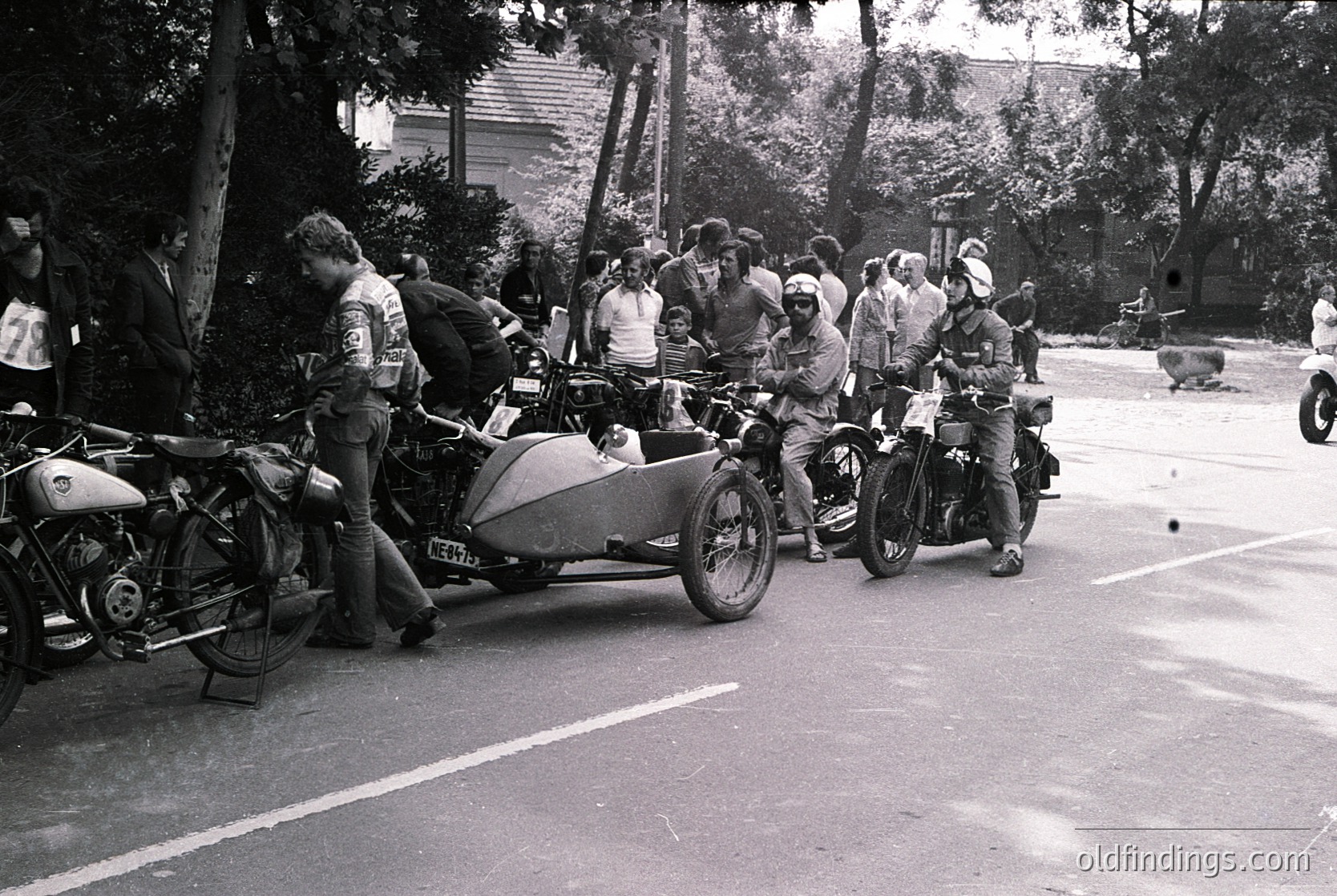 Vintage sidecar motorcycles lined up on a quiet road, surrounded by mid-century attire. Riders in helmets and leather jackets, likely 1960s–1970s. Urban/suburban setting with trees and residential buildings in background.