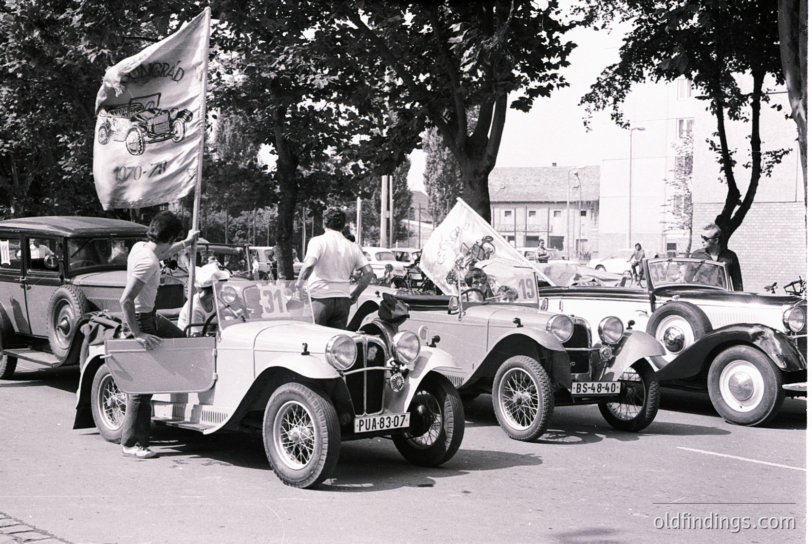 Vintage 1930s road rally with classic cars and vintage flags. Participants wave banners, one featuring a bicycle logo, in a lined street event. Wooden buildings and parked vintage vehicles (, , , , ).