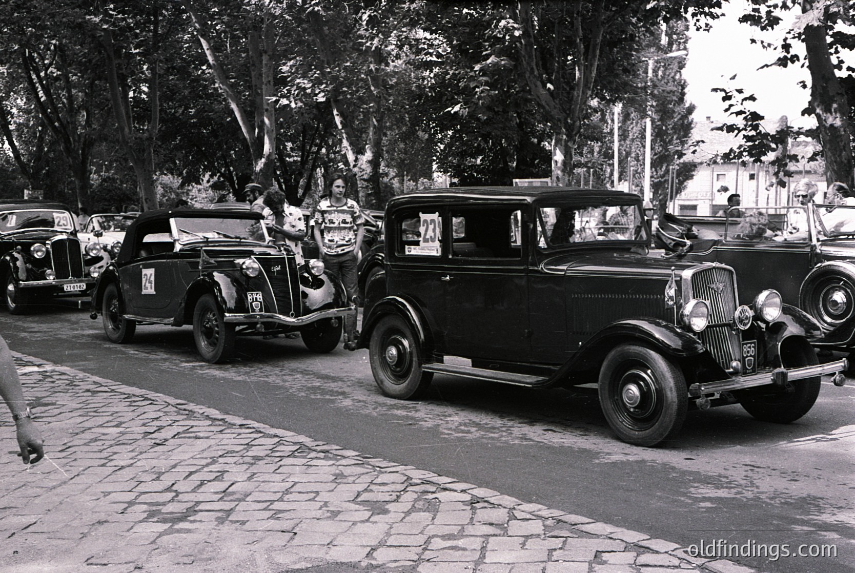 Vintage street scene featuring three classic cars (1930s-1940s) parked on cobblestone pavement. Open-top convertible on left, sedan center, and closed coupe on right. Group of people in mid-20th-century attire posing near vehicles. Lush trees and urban backdrop suggest European city setting.