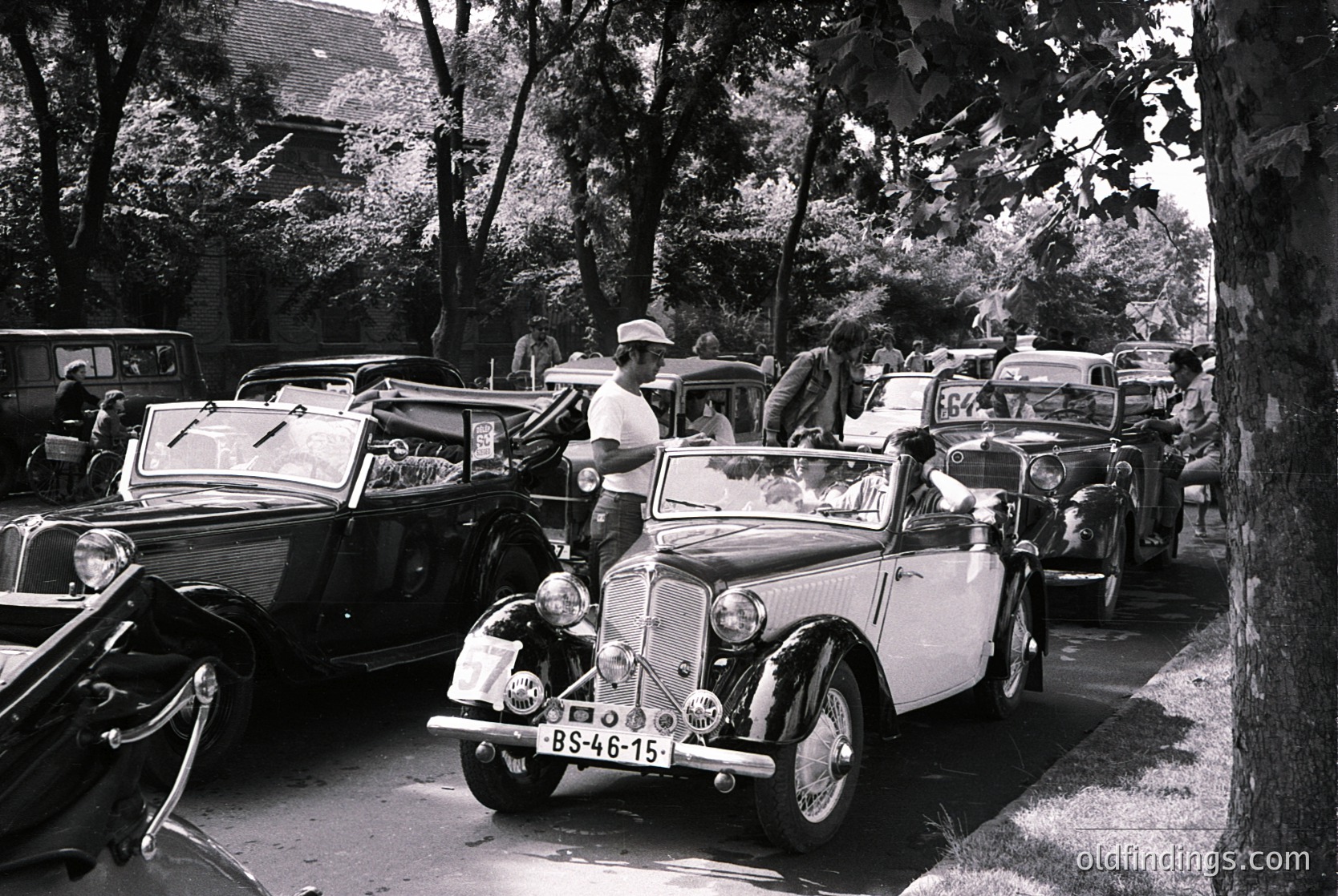 Vintage European roadside gathering featuring classic cars from the 1930s–1950s, including a prominent white convertible with visible license plate "BS-46-15." Shaded by trees, the scene captures mid-century automotive culture, likely in a rural or suburban setting. Crowds in period attire suggest a social or celebratory event.