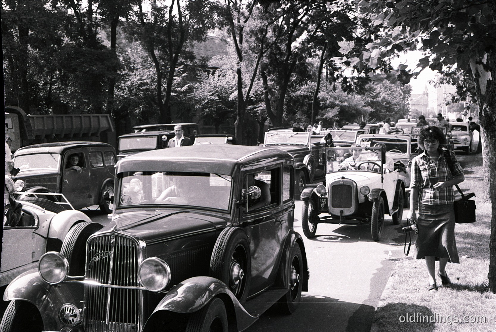 Vintage street scene featuring classic cars from the 1920s–1930s, parked and driving on a tree-lined road. A woman in a long skirt and headscarf walks beside a vintage sedan. Architectural details of a distant building visible in background.