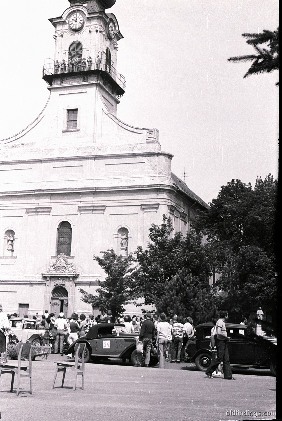 Neoclassical church with prominent clock tower and arched entrance, surrounded by vintage cars and mid-20th century attire. Crowd gathers in courtyard, suggesting a public event or gathering. Likely Eastern European architecture, mid-1950s–1960s.