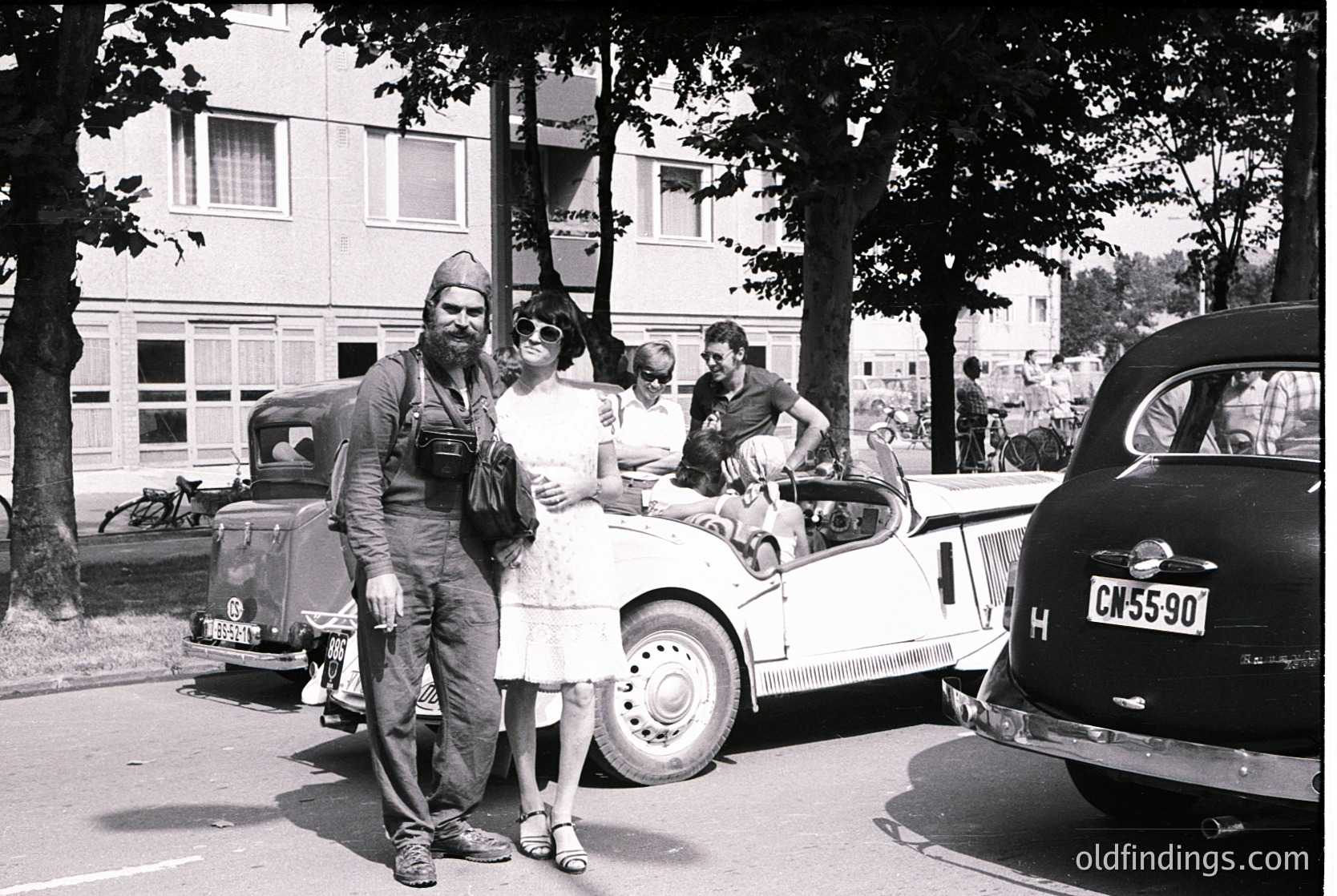 Vintage black-and-white street scene featuring a man in military uniform (likely 1940s–1950s) posing with a woman in a floral dress beside a vintage Volkswagen Beetle (pre-1960s). Surrounding cars include a classic Citroën and a black sedan with visible license plate. Residential buildings with large windows and trees in the background suggest a European urban setting.
