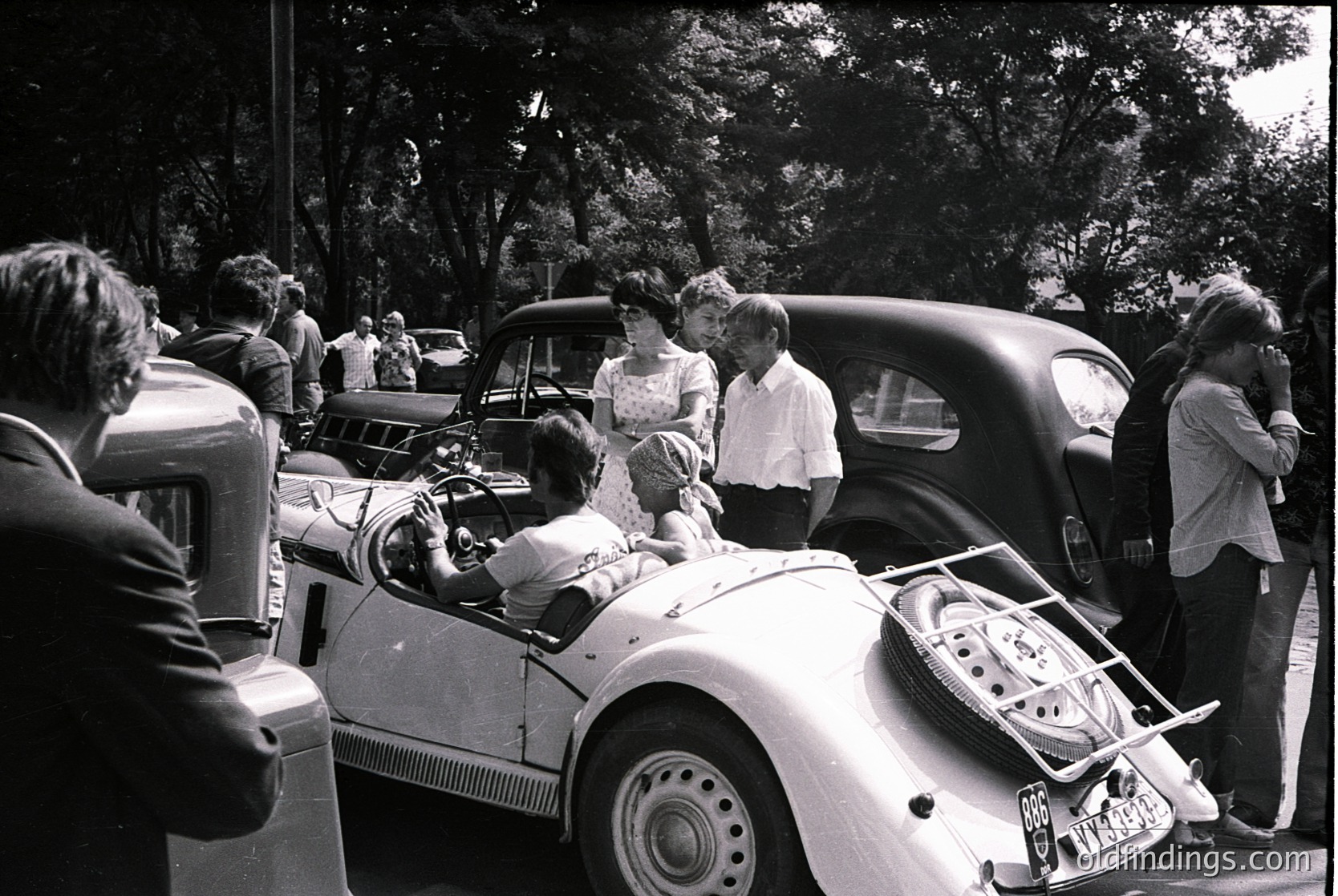 Vintage mid-century roadside gathering featuring classic convertibles and casual 1950s-60s attire. Group of people in light summer dresses and suits pose near a white vintage car with chrome accents and a luggage rack. Lush green trees frame the scene, suggesting a park or suburban setting.