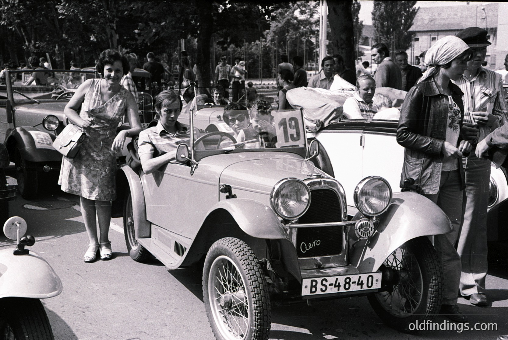 Classic vintage car (BS-48-40) with open-top design and dual headlamps, likely a 1930s–1940s European model, parked in a mid-century street scene. Crowd of casually dressed onlookers, including women in floral dresses and headscarves, observing. Urban setting with trees, parked cars, and residential buildings in background.