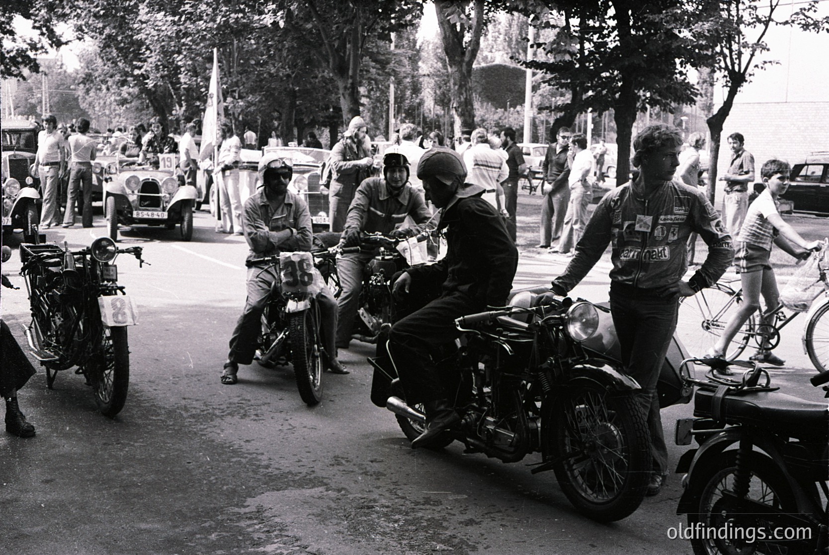 Classic black-and-white street scene featuring vintage motorcyclists in protective gear, likely 1960s–1970s. Riders pose on polished chrome motorcycles with round headlights, surrounded by spectators and parked cars. Urban setting with trees and a crowd, suggesting a public event or rally.