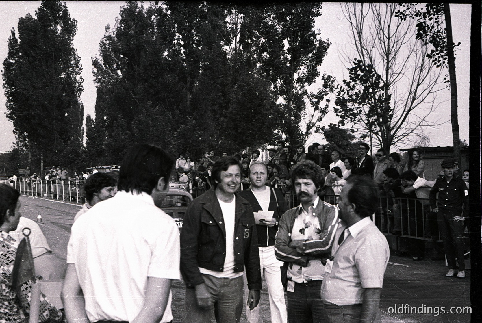 Black-and-white street scene featuring a group of five adults in casual 1970s attire—leather jackets, striped shirts, and bell-bottoms—engaged in conversation. Trees and a metal fence frame the background, suggesting an outdoor public event or festival. Crowds and tents hint at a communal gathering.